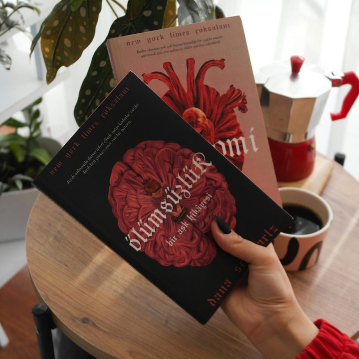 A hand holding two books near a coffee pot and houseplants on a table.